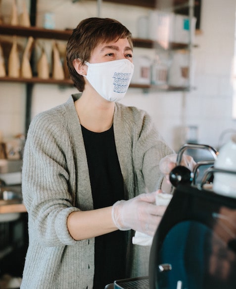 MG Batts smiles under a white mask with "vote" on it in blue while brewing coffee