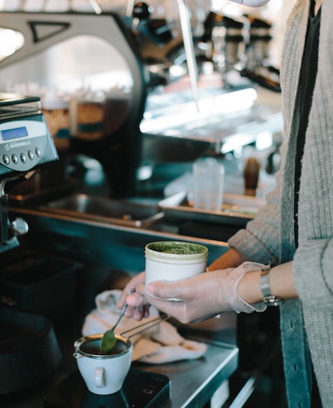 The hands of a person dumping a spoonful of green machta into a tea strainer